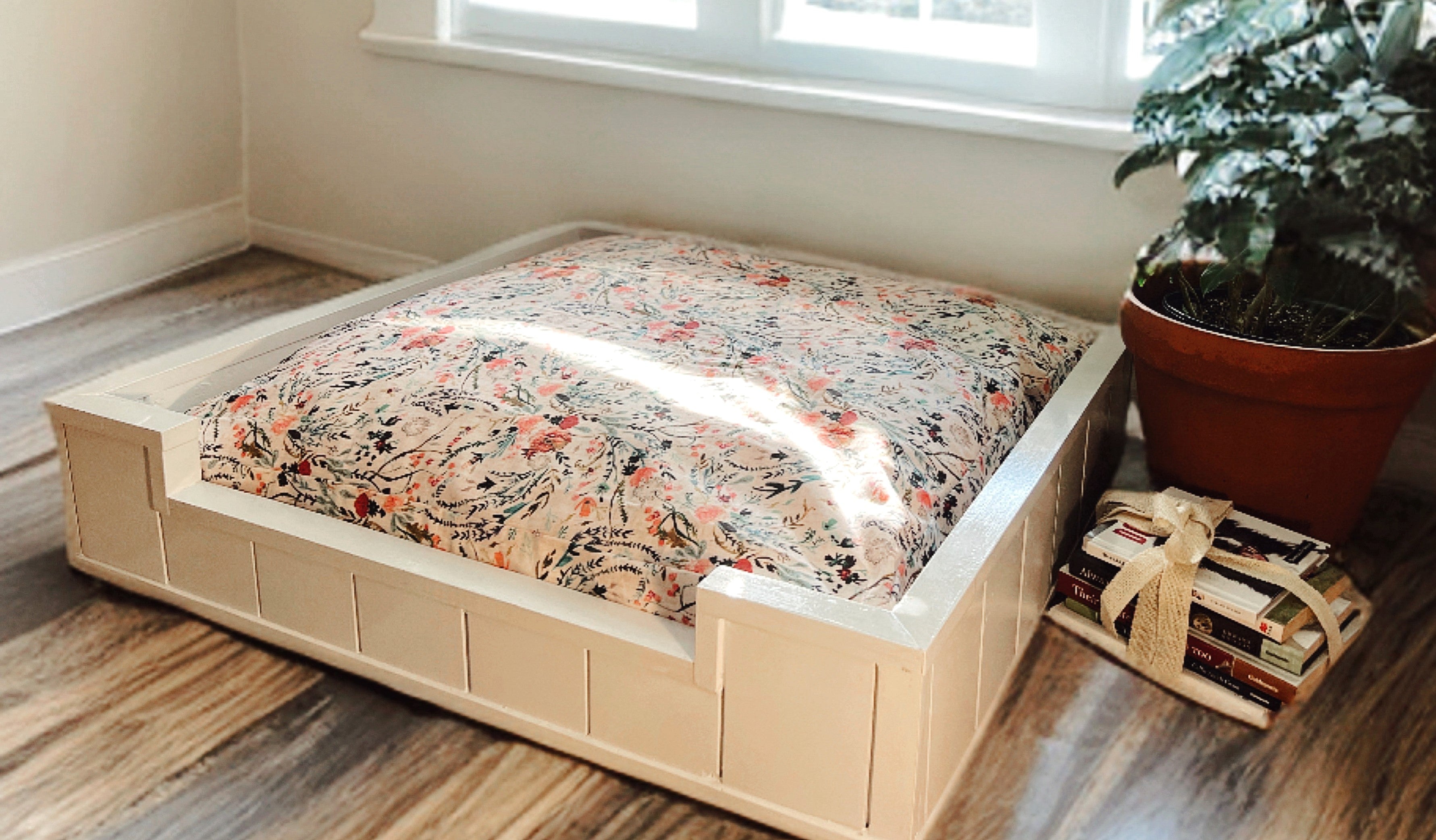 Wooden pet bed with floral cushion on a wooden floor next to a window and plant.