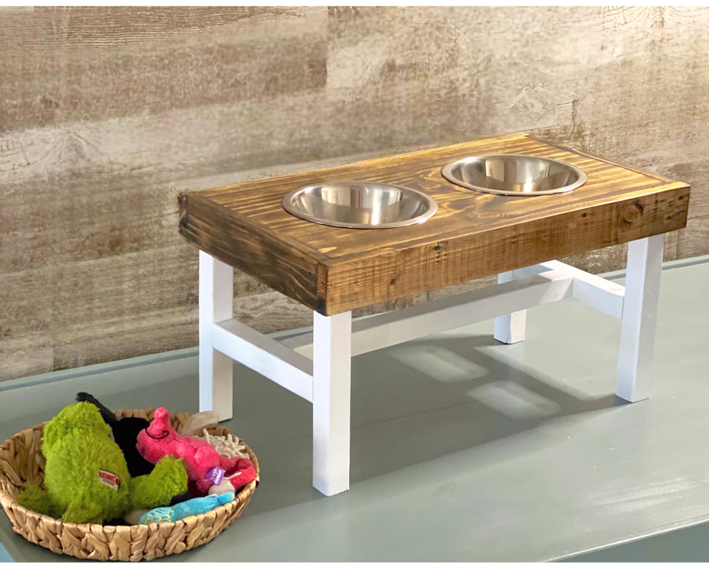 Wooden pet feeder with two bowls on a white surface, next to a basket of toys.Wooden pet feeding station with two bowls on a wooden floor. Raised dog bowl stand, dog bowl holder, large dog bowl stand, small dog bowl stand, wooden dog bowl stand raised, elevated pet bowl stand, elevated dog bowl stand, dog bowl stand wood,